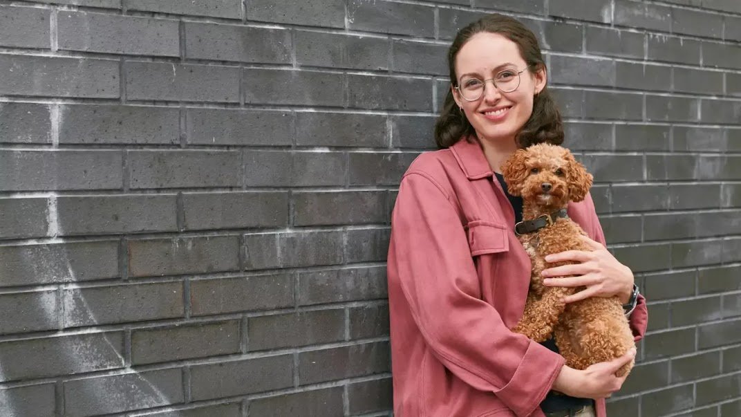 Sylvie wears a pink jacket and leans against a dark grey brick wall. She is smiling and holding a small brown dog